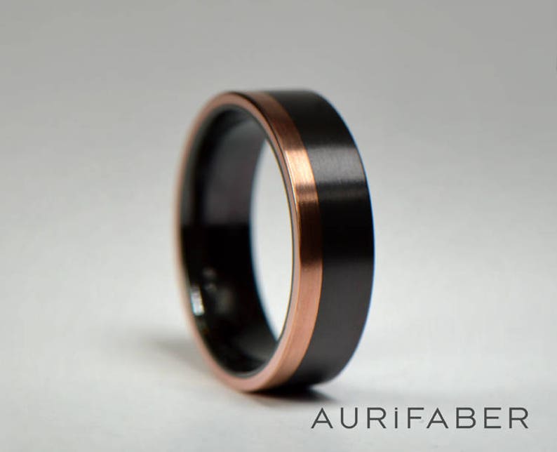 May include: A black and rose gold wedding band with a brushed finish. The band has a thin rose gold inlay around the edge.