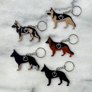 May include: Five German Shepherd dog keychains, each featuring a silver-toned metal split ring and a small dangling heart charm. The keychains display various coat color patterns including black and tan, solid black, and reddish-brown and black.