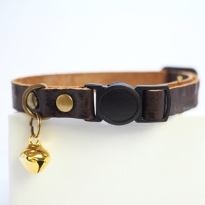 Op de afbeelding: Een bruine leren kattenhalsband met een zwarte plastic gesp en een gouden bel.