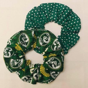 May include: Two green scrunchies. One is patterned with white polka dots, and the other features a repeating design with the words "COLORADO STATE" and a ram logo in white and yellow on a green background. These hair accessories are perfect for showing school spirit.