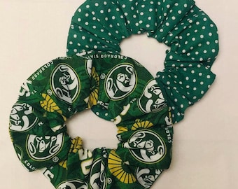 May include: Two green scrunchies. One is patterned with white polka dots, and the other features a repeating design with the words "COLORADO STATE" and a ram logo in white and yellow on a green background. These hair accessories are perfect for showing school spirit.