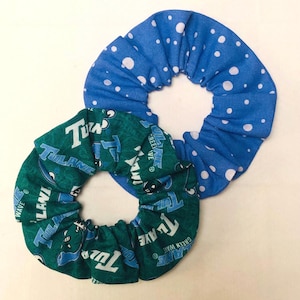 May include: Two scrunchies. One is blue with white polka dots. The other is green with white text and the words "Tulane" and "Green Wave". These hair accessories are perfect for adding a pop of color to any hairstyle.