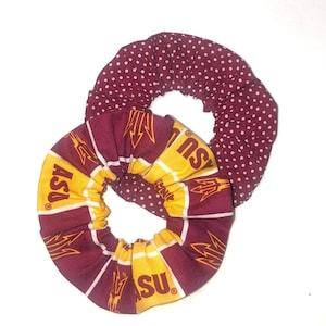 May include: Two scrunchies. One is maroon with white polka dots. The other is maroon and gold with the letters "ASU" and a stylized pitchfork logo. The scrunchies are made of fabric and are circular.