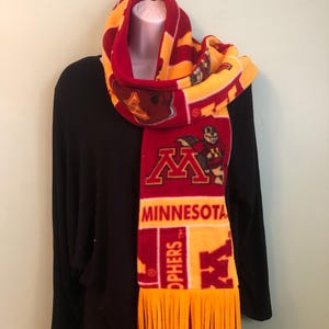 Könnte beinhalten: Ein roter, goldener und kastanienbrauner Fleece-Schal mit dem Wort "MINNESOTA" und dem Team-Maskottchen. Der Schal hat einen Fransen am unteren Ende und wird auf einer Schaufensterpuppe präsentiert. Der Schal ist ein Fan-Accessoire.