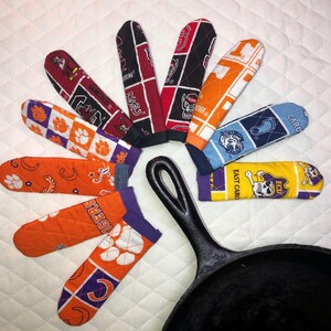May include: A collection of colorful oven mitts and socks with various college team logos and patterns, arranged in a circular fashion around a black cast iron skillet. The mitts and socks feature team colors like orange, purple, red, and blue.