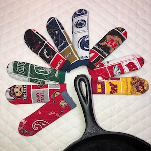 May include: A collection of colorful, patterned pot handle covers and a cast iron skillet. The covers feature college team logos and colors, including Ohio State, Penn State, and Maryland. The covers are arranged around the skillet.