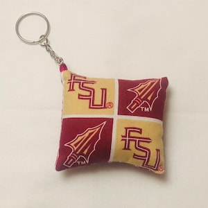 May include: A square keychain featuring a maroon and gold pattern with the letters "FSU" and a spearhead logo. The keychain has a silver metal ring and chain attached to a small, padded fabric square.