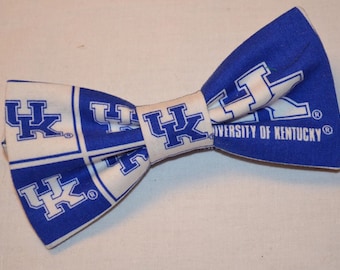 Puede incluir: Una pajarita azul y blanca con un patrón de cuadrícula repetido del logotipo UK de la Universidad de Kentucky. La tela muestra el texto University of Kentucky en letras blancas en el lado derecho de la pajarita con símbolos de marca registrada.