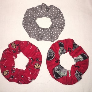 Könnte beinhalten: Draufsicht auf drei Stoff-Haargummis. Eines ist grau mit weißen Punkten. Die anderen beiden sind rot, eines mit Paisley-Muster und das andere mit einem Logo-Design. Diese Haaraccessoires sind perfekt, um einen Farbtupfer zu setzen.