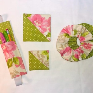 Puede incluir: Conjunto de accesorios de tela hechos a mano con un estampado floral. Los artículos incluyen un portalápices, posavasos y una scrunchie. El estampado presenta flores rosas y hojas verdes sobre un fondo crema, con lunares verdes. El portalápices tiene un lazo elástico blanco.