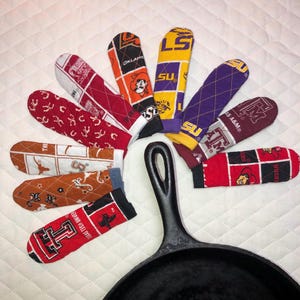 May include: A selection of patterned pot handle covers, each with collegiate team logos and designs. The covers are arranged around a black cast iron skillet, displaying a variety of team colours and patterns.