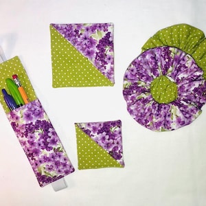 Puede incluir: Un conjunto de artículos de tela hechos a mano con un estampado floral y de lunares. El conjunto incluye un portalápices con bolígrafos, dos posavasos cuadrados y un artículo redondo, todo en tonos morados, verdes y blancos.