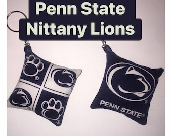 May include: Two Penn State Nittany Lions keychains. One keychain features a paw print and the Penn State logo in a white and navy blue pattern. The other keychain has the Penn State logo and text on a navy blue background.