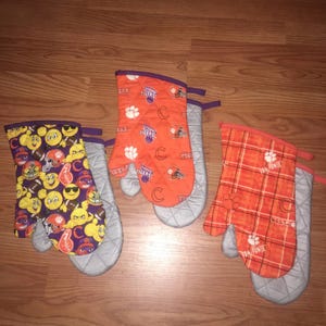 May include: Three oven mitts with quilted grey linings. The left mitt features yellow emojis and footballs on a purple background. The centre mitt is orange with repeating Tigers logos and paw prints. The right mitt has an orange plaid pattern.