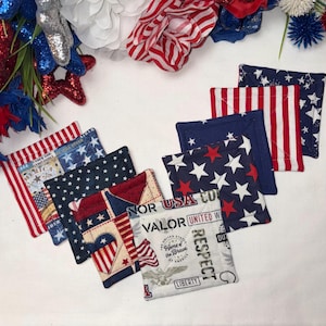 May include: A collection of square fabric coasters featuring red, white, and blue patterns including stars, stripes, and text such as VALOR, USA, RESPECT, and LIBERTY. The quilted coasters are arranged in a cluster with visible stitching details.