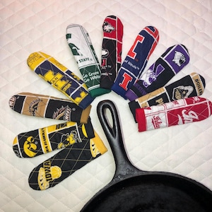 Op de afbeelding: Een verzameling gewatteerde handgreepbeschermers voor gietijzeren koekenpannen in verschillende kleuren en patronen, inclusief universiteitslogo's en teamnamen. De hoezen zijn gerangschikt rond een zwarte gietijzeren koekenpan.