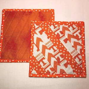 May include: Two square quilted pot holders. One is orange with a diagonal pattern. The other is white with orange chevrons and the text "UNIVERSITY OF". Both have orange polka dot borders and are likely handmade.