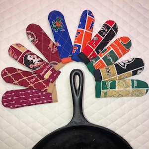 May include: A black cast iron skillet with a handle, surrounded by several colorful oven mitts. The mitts feature various college team logos and patterns in maroon, blue, orange, green, and black. The mitts are made of quilted fabric.