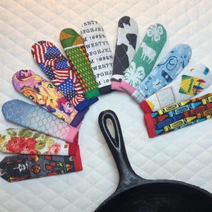 Könnte beinhalten: Eine Sammlung bunter Topflappen mit verschiedenen Mustern, darunter amerikanische Flaggen, Blumendesigns und Zeichentrickfiguren. Die Topflappen sind um eine schwarze gusseiserne Pfanne angeordnet, was ein Küchenutensilien-Thema zeigt.