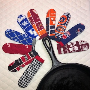 Könnte beinhalten: Eine Sammlung von Ofenhandschuhen und Topflappen in verschiedenen Farben und Mustern, darunter Teamlogos und Hahnentrittmuster. Eine schwarze gusseiserne Bratpfanne befindet sich im Vordergrund. Die Handschuhe und Halter sind um die Pfanne angeordnet.