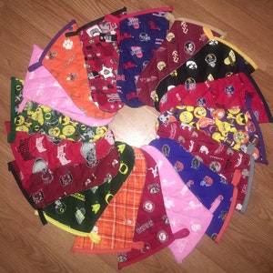 May include: A collection of quilted oven mitts in various colors and patterns, including sports team logos and university emblems. The mitts are arranged in a circular pattern on a wooden surface. Each mitt has a loop for hanging.