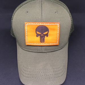 Könnte beinhalten: Eine grüne und schwarze Mesh-Baseballkappe mit einem braunen Lederpatch, auf dem ein schwarzer Totenkopf mit gekreuzten Knochen abgebildet ist.