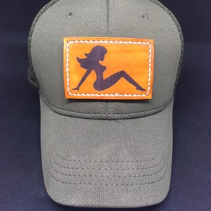 Puede incluir: Una gorra de camionero verde con un parche de cuero marrón que presenta la silueta de una mujer. El parche tiene costuras blancas alrededor de los bordes.
