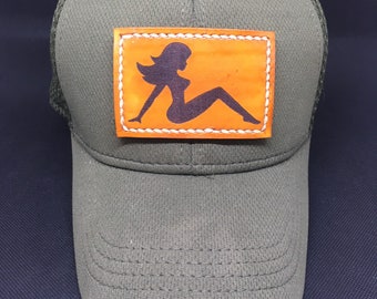 May include: A green trucker hat with a brown leather patch featuring a silhouette of a woman. The patch has white stitching around the edges.