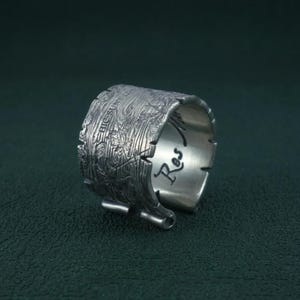 Op de afbeelding: Een zilveren ring met een getextureerd, houtnerfachtig oppervlak. De ring heeft een uniek, gescheurd papierontwerp met het woord "Res" aan de binnenkant gegraveerd. Het buitenoppervlak heeft geëtste details en de tekst "HIC SUNT DRACONIS".