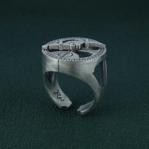 Può includere: Un anello argentato con un design meccanico dettagliato sulla parte superiore. L'anello presenta un motivo circolare con un elemento centrale e una fascia strutturata. Il design aperto consente la regolazione delle dimensioni. L'anello è su uno sfondo verde scuro.