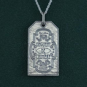 Peut inclure: Collier en argent avec un pendentif en forme d'étiquette qui porte l'inscription "Onguent de Chimere" avec un motif floral. L'étiquette a une surface texturée.