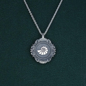 May include: A silver necklace with a detailed pendant featuring a white seashell design. The pendant is framed by a decorative border.