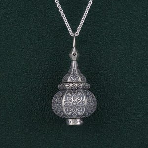 Könnte beinhalten: Eine silberne Halskette mit einem detaillierten, verzierten Anhänger. Der Anhänger ist in Form eines kleinen, kuppelförmigen Gebäudes mit einer spitzen Spitze gestaltet. Der Anhänger hat einen Klappdeckel und ist mit dem Wort "Mina" graviert.