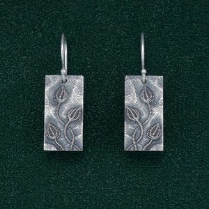 May include: A pair of silver earrings with a textured rectangular design featuring a stylized floral motif.