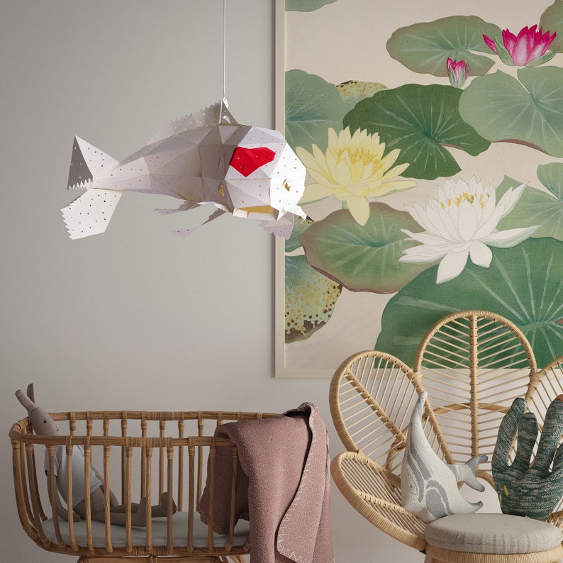 K&ouml;nnte beinhalten: Eine wei&szlig;e, fischf&ouml;rmige Lampe aus Papier mit einem roten Herzdetail h&auml;ngt von der Decke &uuml;ber einem Weidenkorb und einem Weidenstuhl mit einem grauen Wal-Kissen und einem gr&uuml;nen Kaktus-Kissen.
