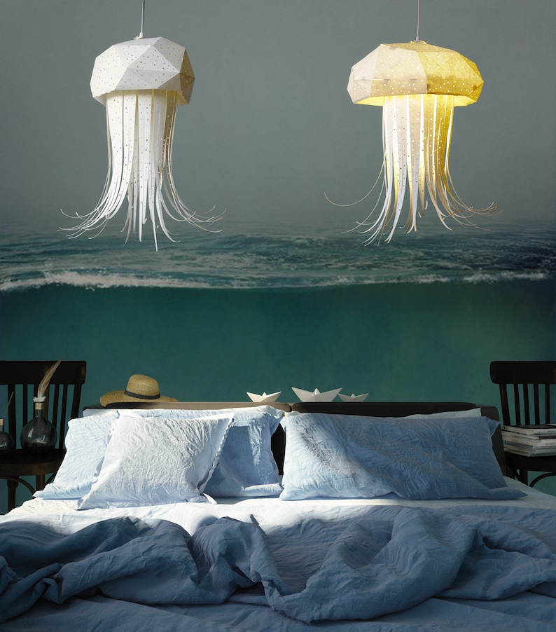 K&ouml;nnte beinhalten: Zwei wei&szlig;e Papier-Medusen-Pendelleuchten h&auml;ngen &uuml;ber einem blauen Bett mit wei&szlig;en Kissen. Der Hintergrund ist ein Wandbild mit einer Ozeanszene.