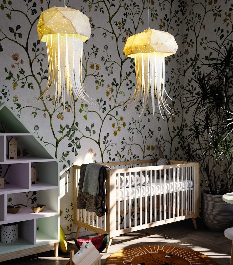 K&ouml;nnte beinhalten: Zwei wei&szlig;e und gelbe Papier-Quallen-Leuchten h&auml;ngen an der Decke eines Kinderzimmers. Der Raum hat eine Tapeten mit Blumenmuster und ein wei&szlig;es Babybett mit einer wei&szlig;en Matratze und einer braunen Decke.