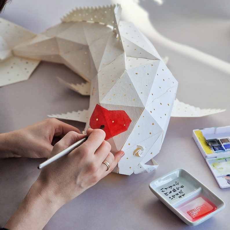 K&ouml;nnte beinhalten: Eine Person bemalt eine wei&szlig;e Papierfischskulptur mit roter Wasserfarbe. Der Fisch hat ein geometrisches Design und ist aus Papier gefertigt. Die Person verwendet einen kleinen Pinsel, um den Fisch zu bemalen. Im Hintergrund befinden sich eine kleine Schale mit Wasser und eine Wasserfarbenpalette.