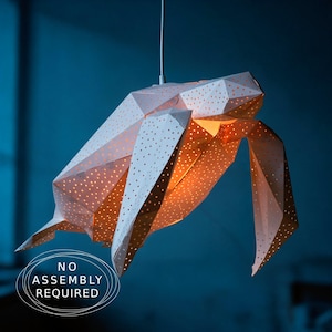 May include: A geometric sea turtle pendant light fixture made of white material with numerous small circular perforations that emit a warm orange glow from within. The structure is suspended by a single cord and features the text NO ASSEMBLY REQUIRED.