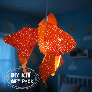 May include: A hanging pendant lamp shaped like a goldfish, constructed from orange geometric panels with a perforated polka dot pattern that allows light to shine through. The lamp is suspended by a white cord and features the text DIY KIT GIFT PACK.
