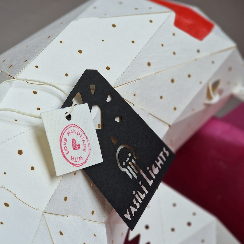 K&ouml;nnte beinhalten: Wei&szlig;e Papierlaterne mit goldenen Punkten und einem schwarzen Etikett mit der Aufschrift "Vasilii Lights" und einem kleinen Herzen mit den Worten "Love with Handmade" in einem roten Kreis.