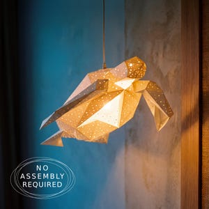 May include: A white geometric sea turtle pendant light fixture with a perforated shell design that emits a warm glow. The turtle is suspended from a single cord and features the text NO ASSEMBLY REQUIRED printed in the lower left corner.
