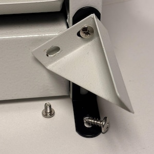 May include: Close-up of white metal corner brackets and screws. The brackets are L-shaped with pre-drilled holes for screws. One screw is loose, and others are attached to the brackets and a black hinge. The hardware is for furniture or construction.