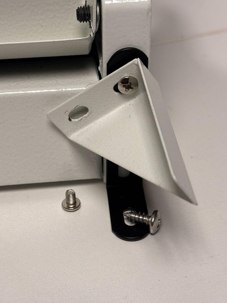 May include: Close-up of white metal corner brackets and screws. The brackets are L-shaped with pre-drilled holes for screws. One screw is loose, and others are attached to the brackets and a black hinge. The hardware is for furniture or construction.