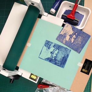 May include: A printmaking setup with a teal-colored roller, a white press, and a blue ink roller. Two blue prints are displayed on a light blue paper. A tube of blue paint and a red handled ink roller are also visible.