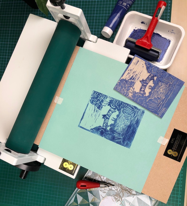 May include: A printmaking setup with a teal-colored roller, a white press, and a blue ink roller. Two blue prints are displayed on a light blue paper. A tube of blue paint and a red handled ink roller are also visible.