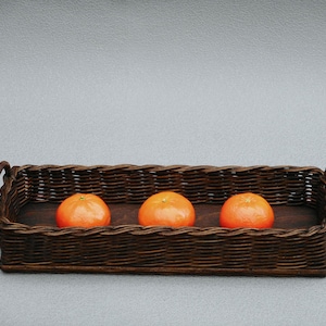 May include: A rectangular brown wicker basket with three bright orange tangerines. The basket has a dark wooden base and curved handles on each end. The tangerines are centered in the basket.