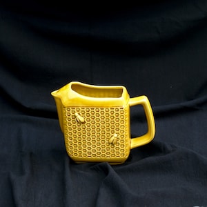 Könnte beinhalten: Gelbe Keramikkrug mit Wabenmuster und zwei Bienenmotiven. Der Krug hat eine rechteckige Form, einen Henkel und einen Ausguss. Der Hintergrund ist ein dunkler Stoff.