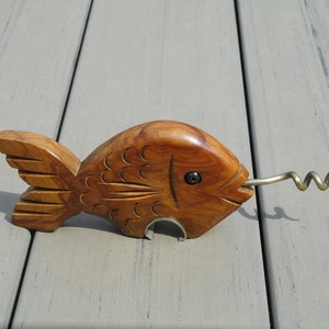 May include: A wooden fish-shaped corkscrew with a metal corkscrew and bottle opener. The fish is carved from light brown wood, with etched details for scales and fins. The corkscrew extends from the fish's mouth.