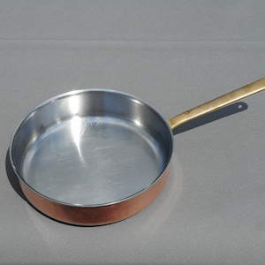 May include: A copper-bottomed frying pan with a stainless steel interior and a brass handle. The pan is round and has a long handle with a hole at the end for hanging. The pan is designed for cooking.